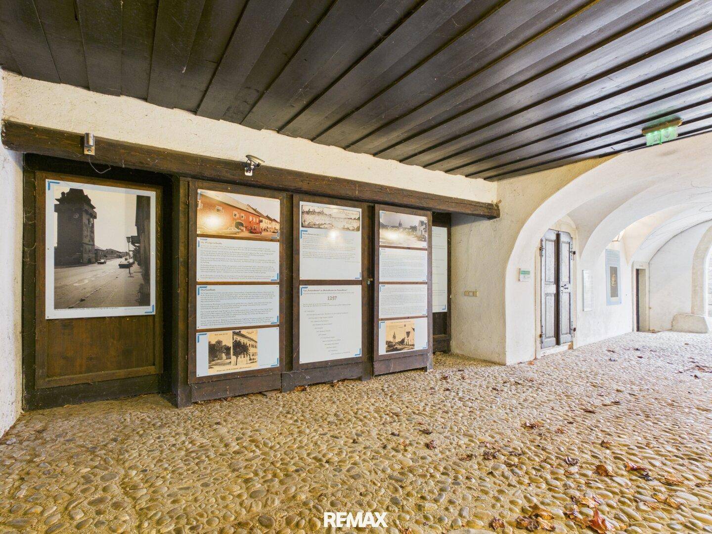 Historischer Durchgang mit Holzbalkendecke, Kieselsteinboden und Ausstellungen an den Wänden.