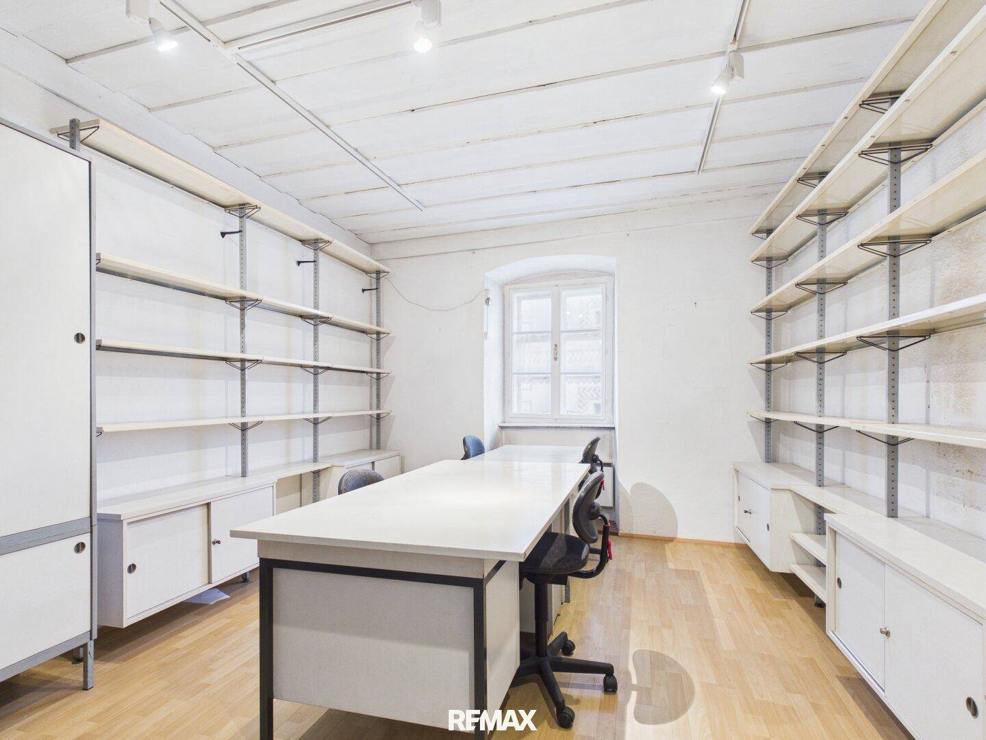 Helles Büro mit Holzboden, Regalsystemen und einem großen Schreibtisch, gut beleuchtet durch ein Fenster.