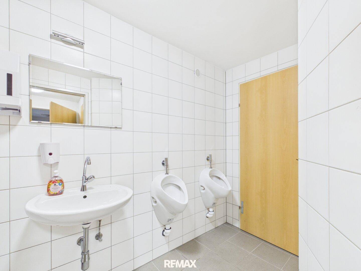 Badezimmer mit Waschbecken, zwei Urinalen und weißen Fliesen, praktisch und hygienisch.