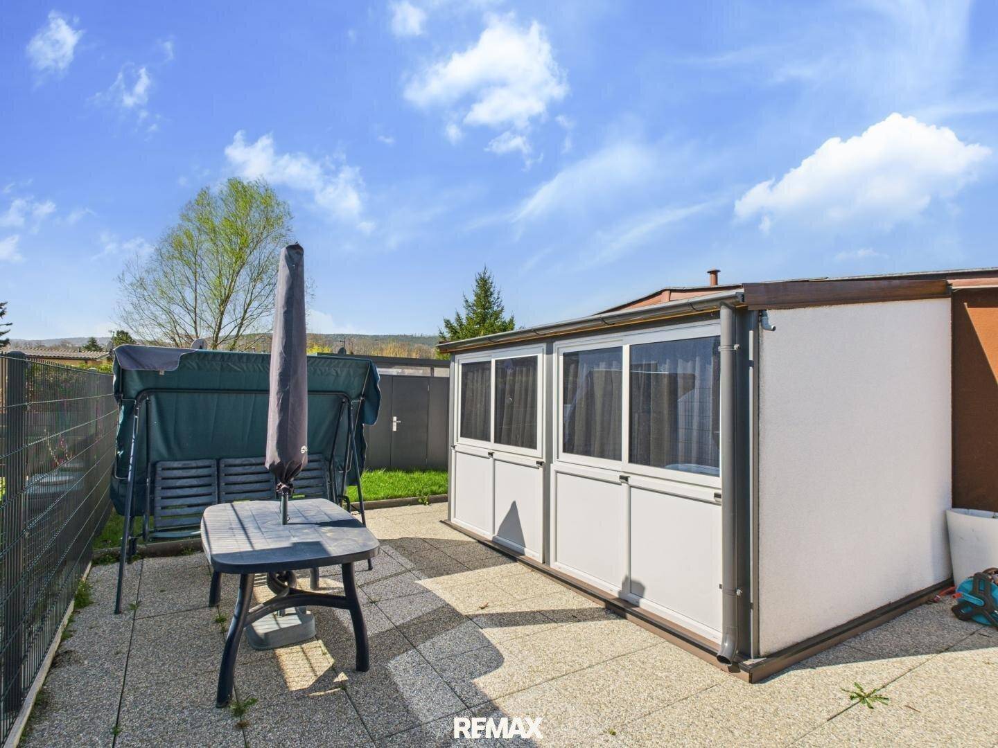 Gepflasterte Terrasse mit Gartenmöbeln, Schaukel und Sonnenschirm neben dem Wintergarten.