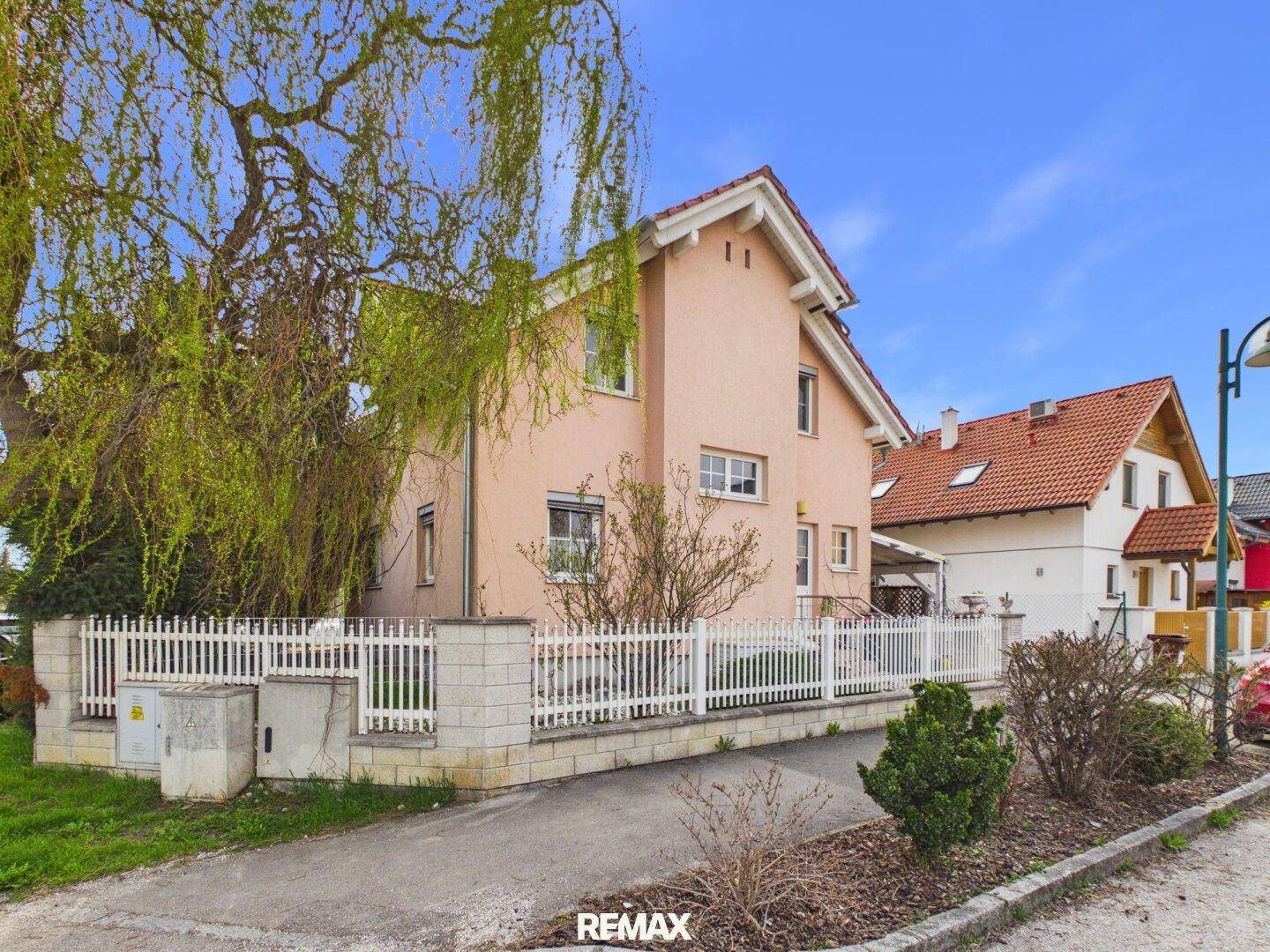 Einfamilienhaus mit rosa Fassade, weißem Zaun und Bäumen im Vorgarten an einem sonnigen Tag.