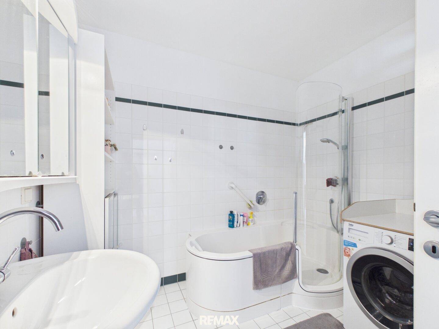 Helles Badezimmer mit Badewanne, Duschkabine, Waschmaschine und weiß-grünen Fliesen.