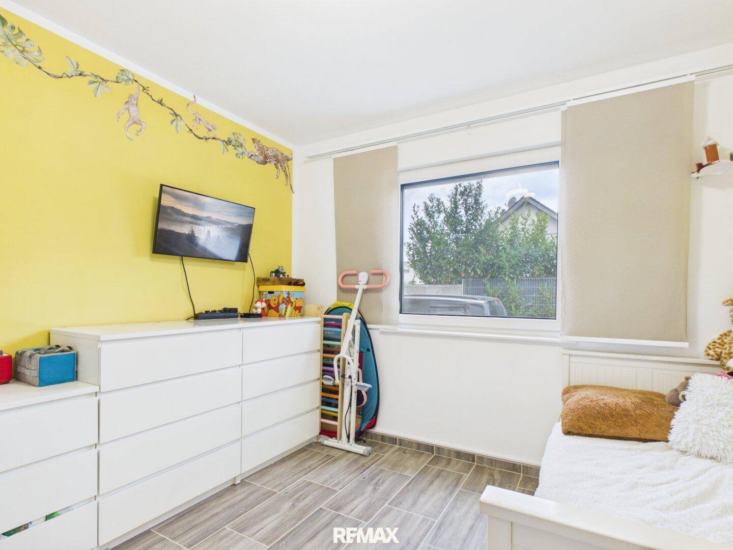 Kinderzimmer mit gelber Akzentwand, weißer Kommode und Blick ins Grüne.
