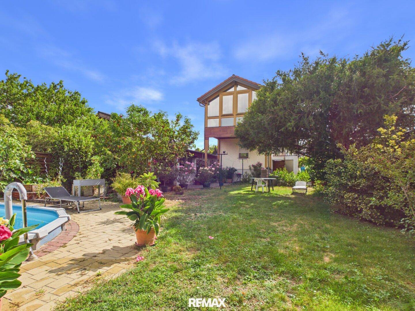 Weitläufiger Garten mit Rasenfläche, Bäumen und einem Haus im Hintergrund unter blauem Himmel.