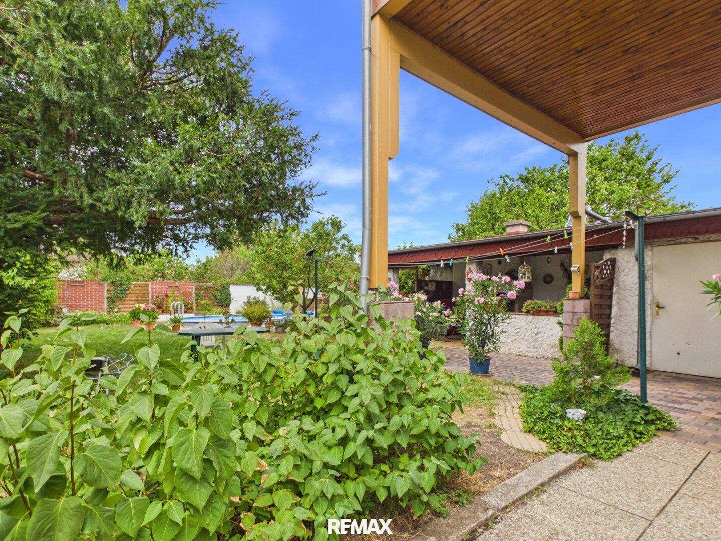 Gartenansicht mit üppiger Bepflanzung und Blick auf die überdachte Terrasse und den Poolbereich.