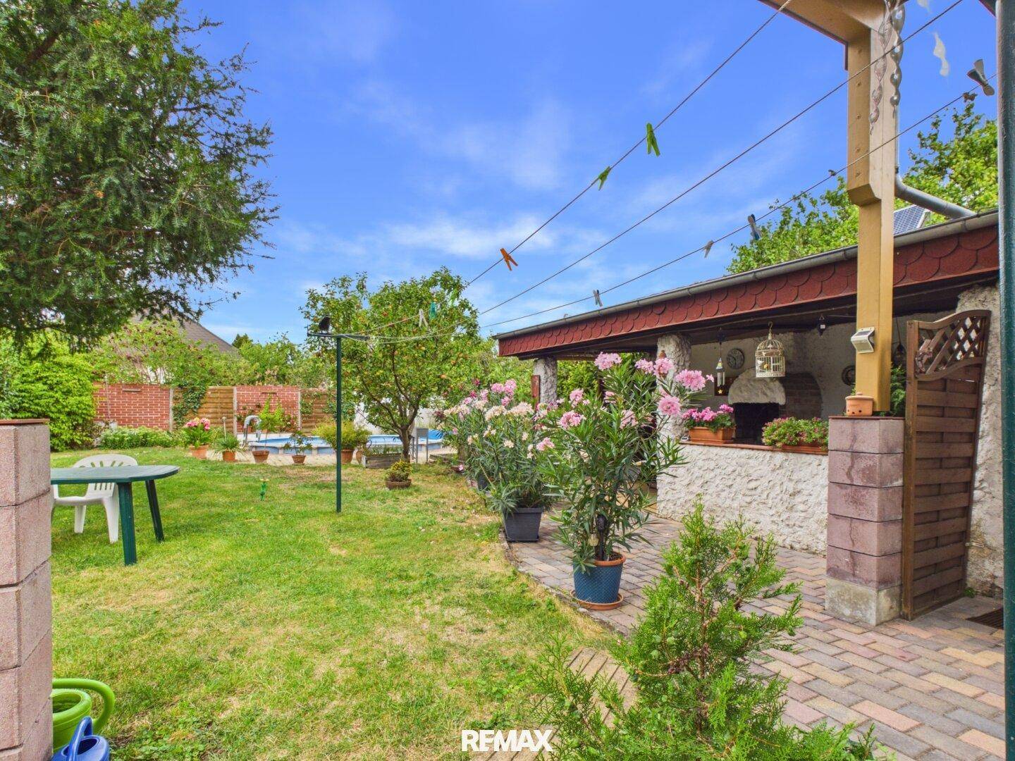 Garten mit Rasen und Blick auf die überdachte Terrasse, geschmückt mit blühenden Pflanzen und Wäscheleinen.
