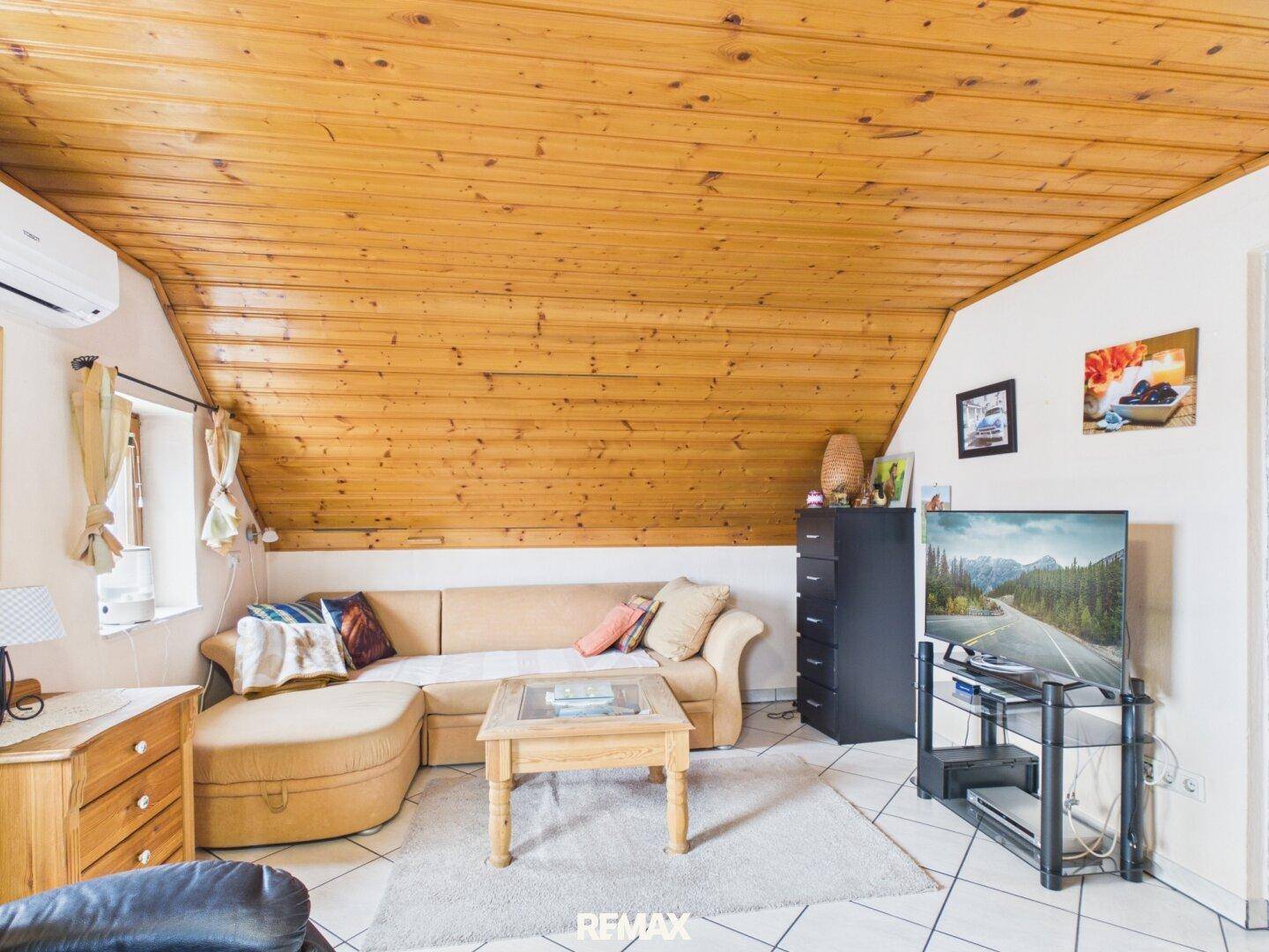 Gemütliches Wohnzimmer im Dachgeschoss mit Sofa, Fernseher und Holzdeckenverkleidung.