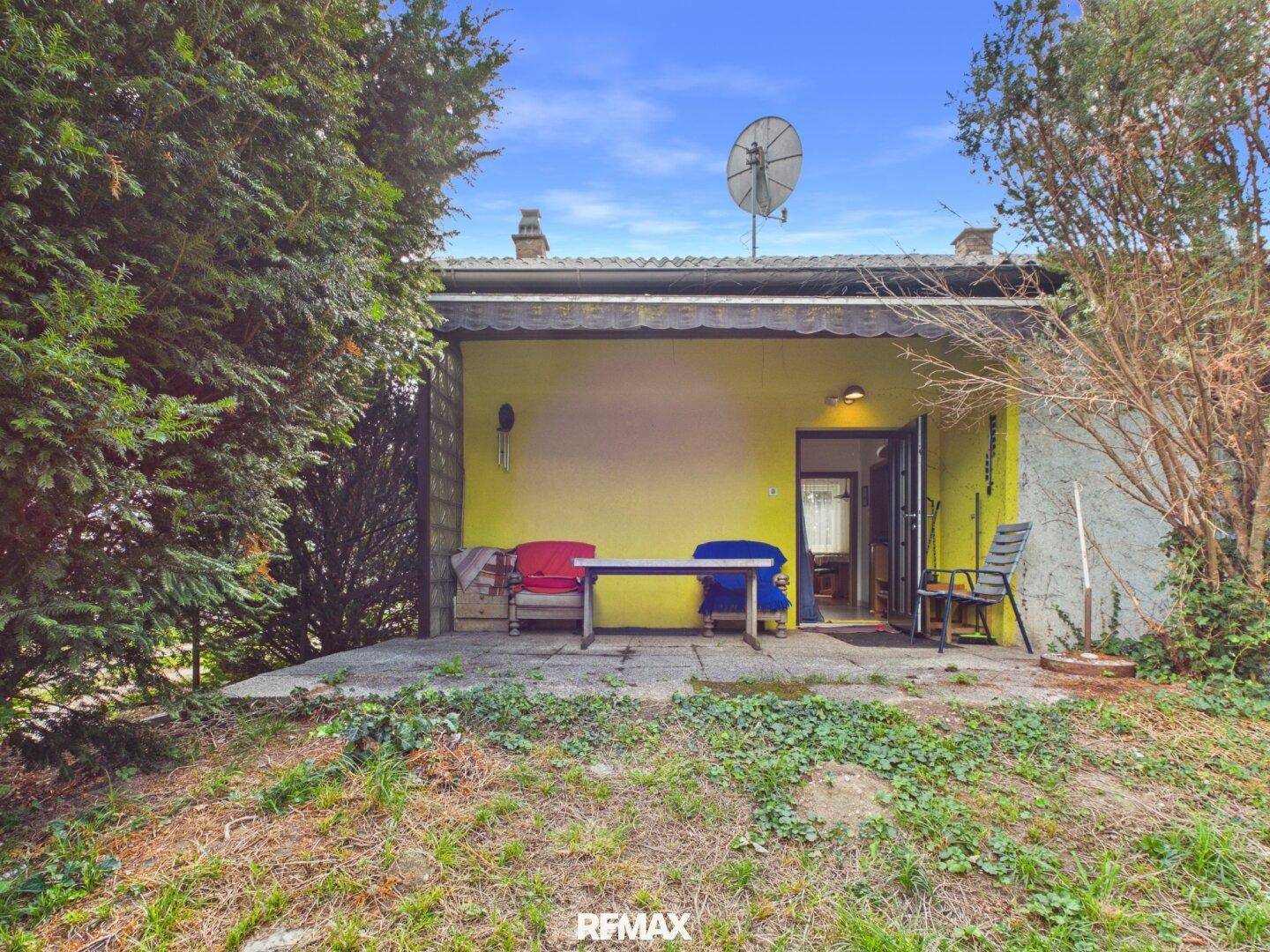 Überdachte Terrasse mit gelber Wand und einfacher Sitzgelegenheit im Freien.