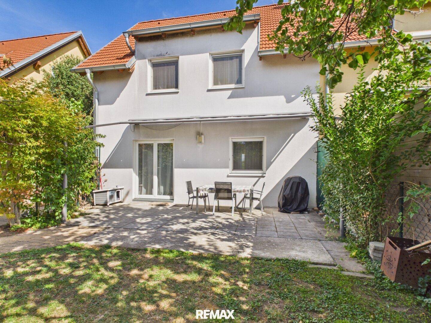 Rückseite des Hauses mit gepflasterter Terrasse, Gartenmöbeln und Rasenfläche unter blauem Himmel.