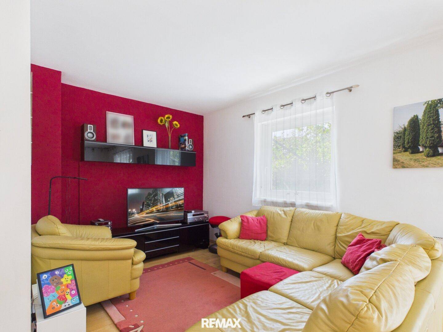 Wohnzimmer mit gelbem Ledersofa, roter Akzentwand und Fenster mit Blick ins Grüne.