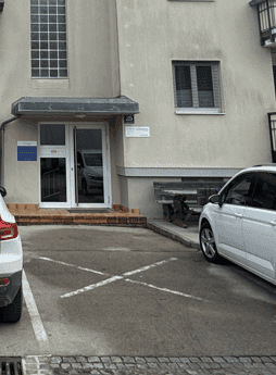 Außenansicht eines Gebäudes mit Eingangsbereich und Parkplätzen davor, schlichte Architektur.