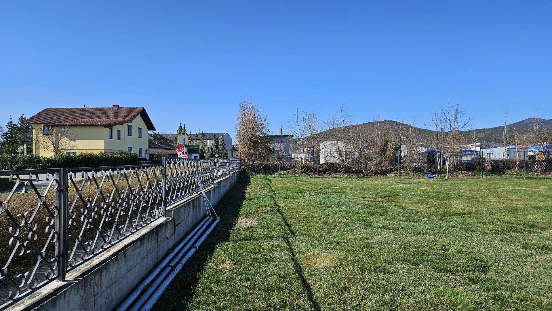 Großes unbebautes Grundstück mit Zaun und angrenzenden Wohnhäusern unter klarem blauen Himmel.