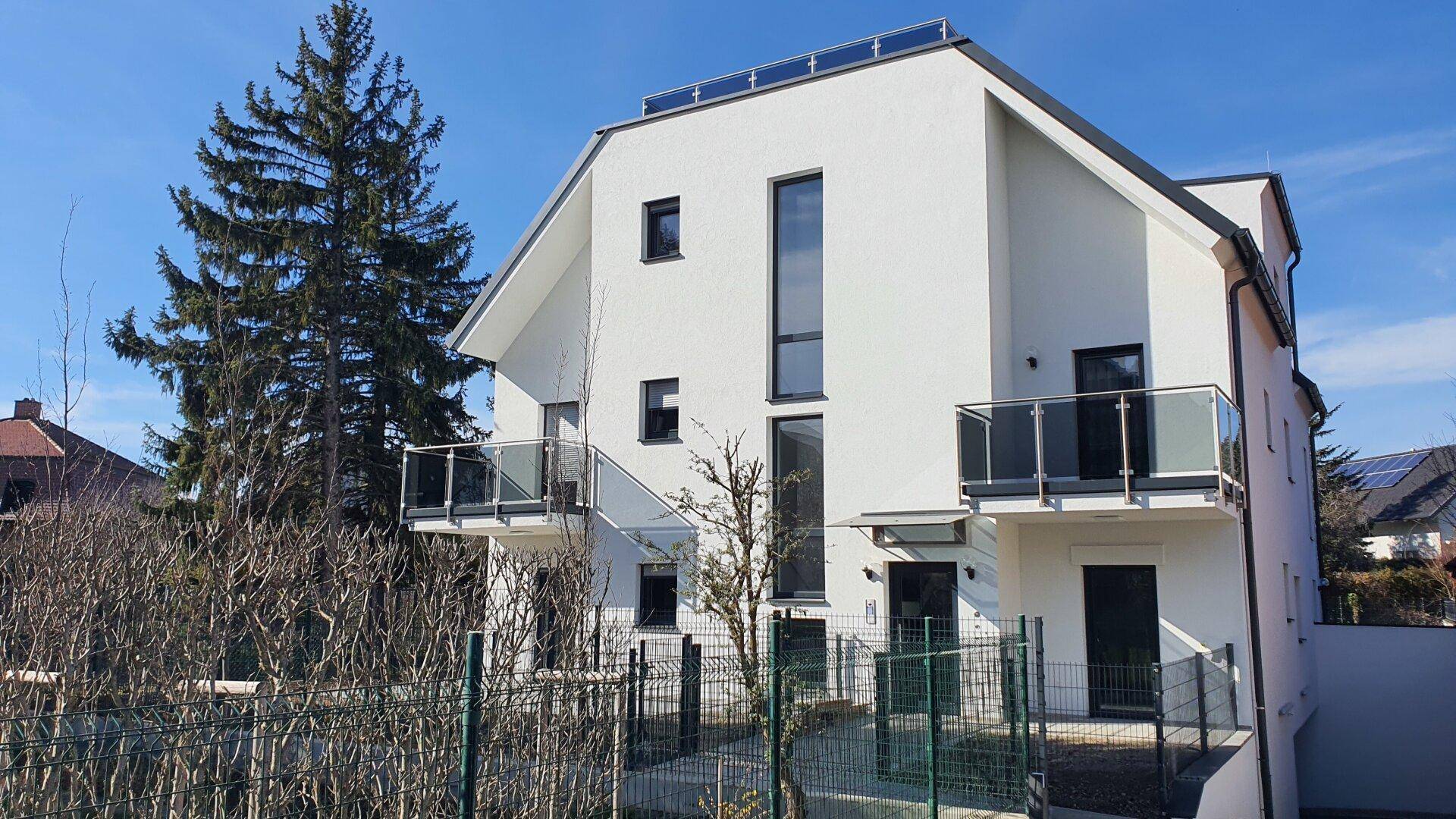 Moderne weiße Fassade eines Neubaus mit Balkonen und Solarpaneelen auf dem Dach.