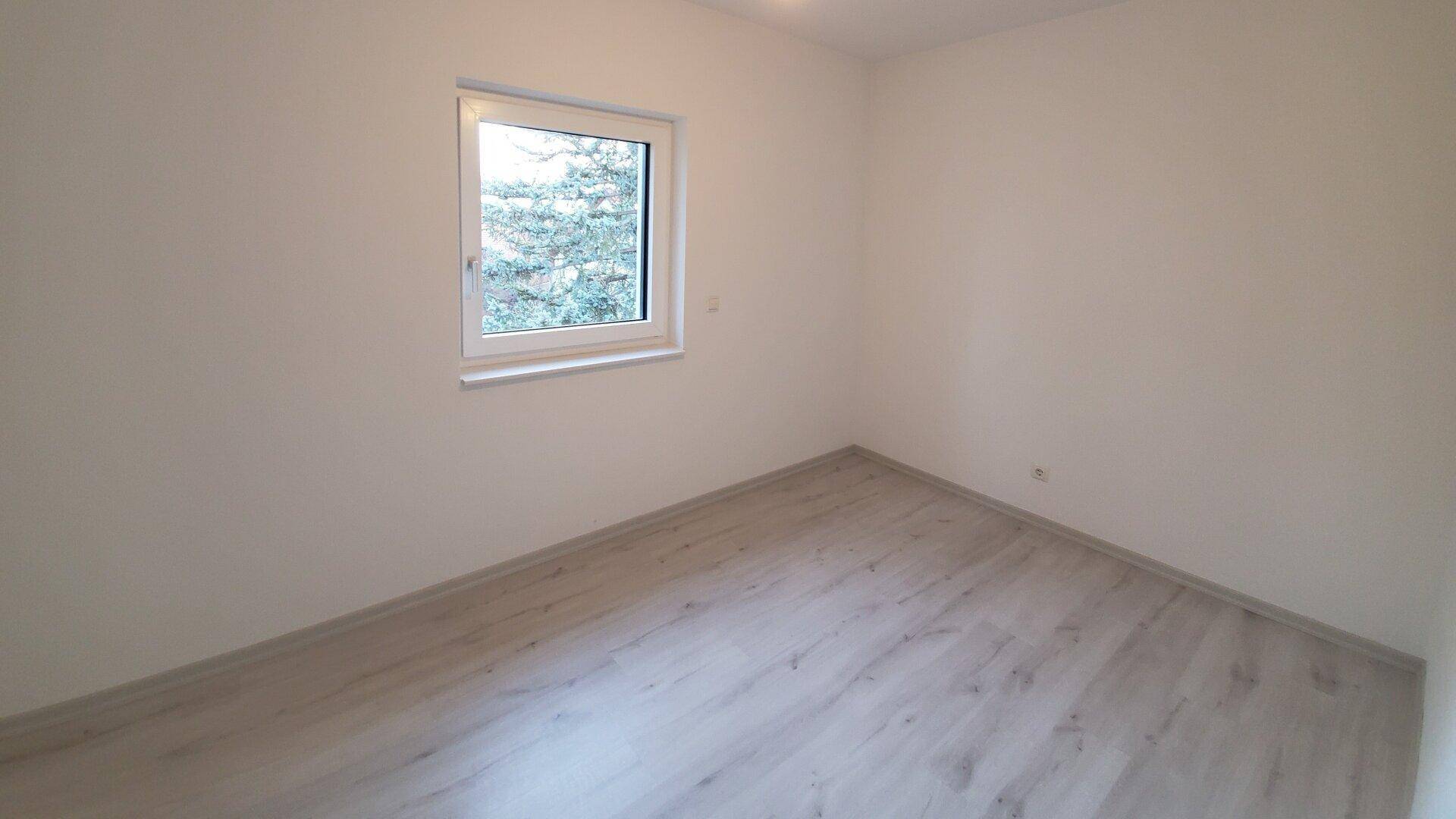 Leerer Raum mit einem Fenster und hellem Laminatboden, bereit zur individuellen Gestaltung.