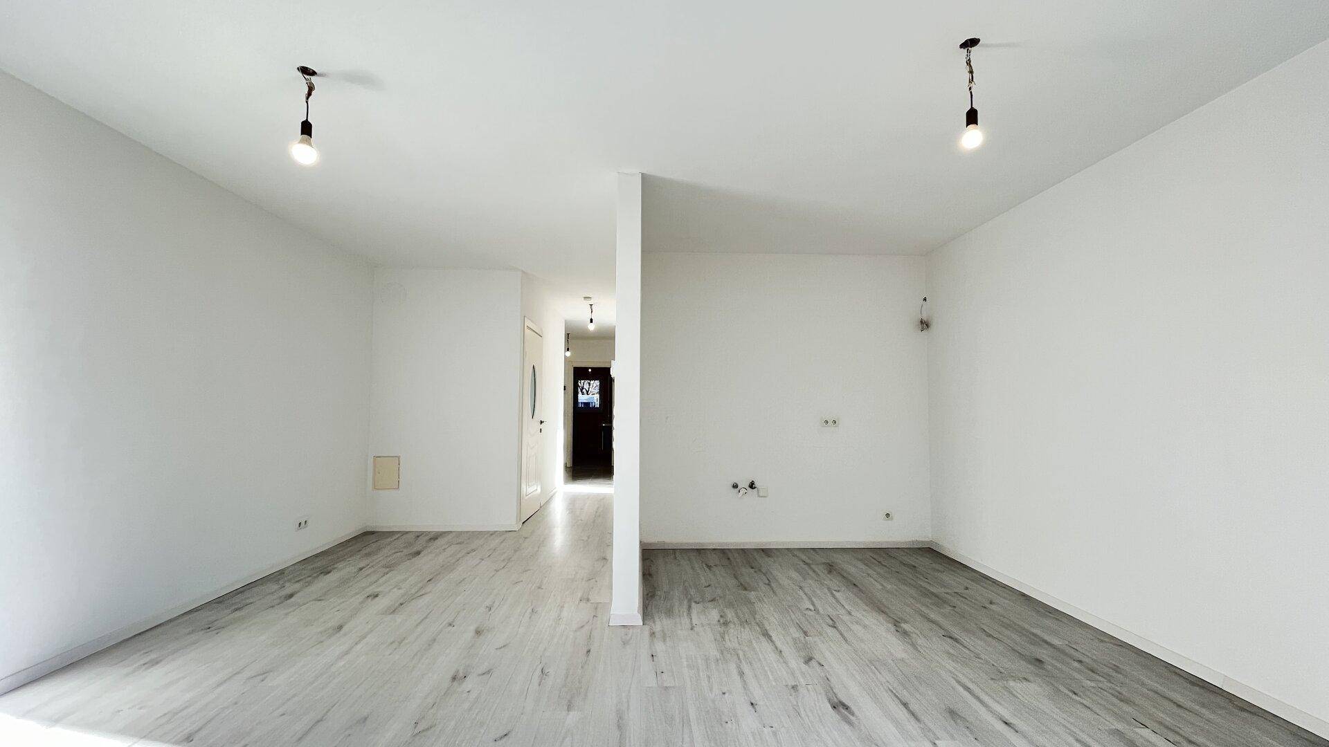 Leerer Wohnraum mit hellem Laminatboden und Blick in den angrenzenden Flur.