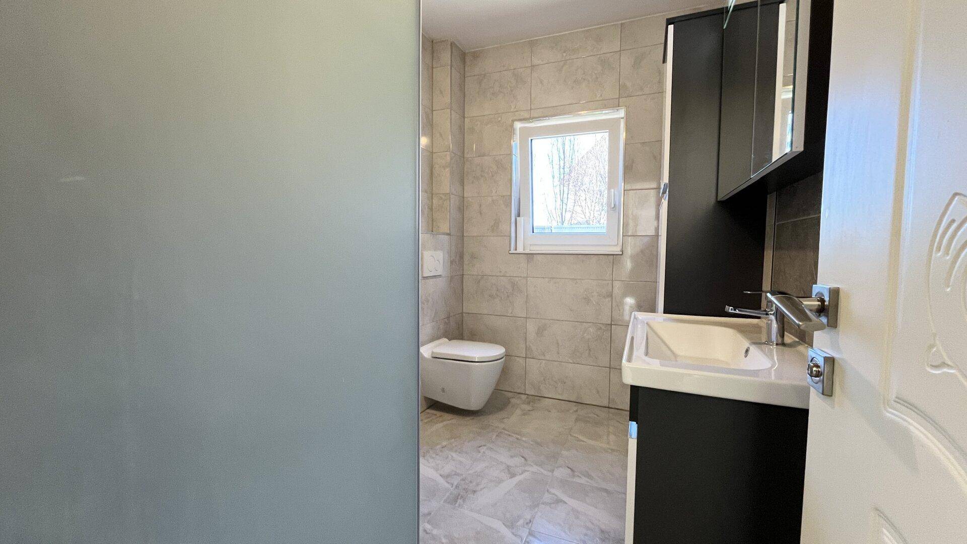 Badezimmer mit Wand-WC, Waschtisch und Fenster für natürliche Belüftung.