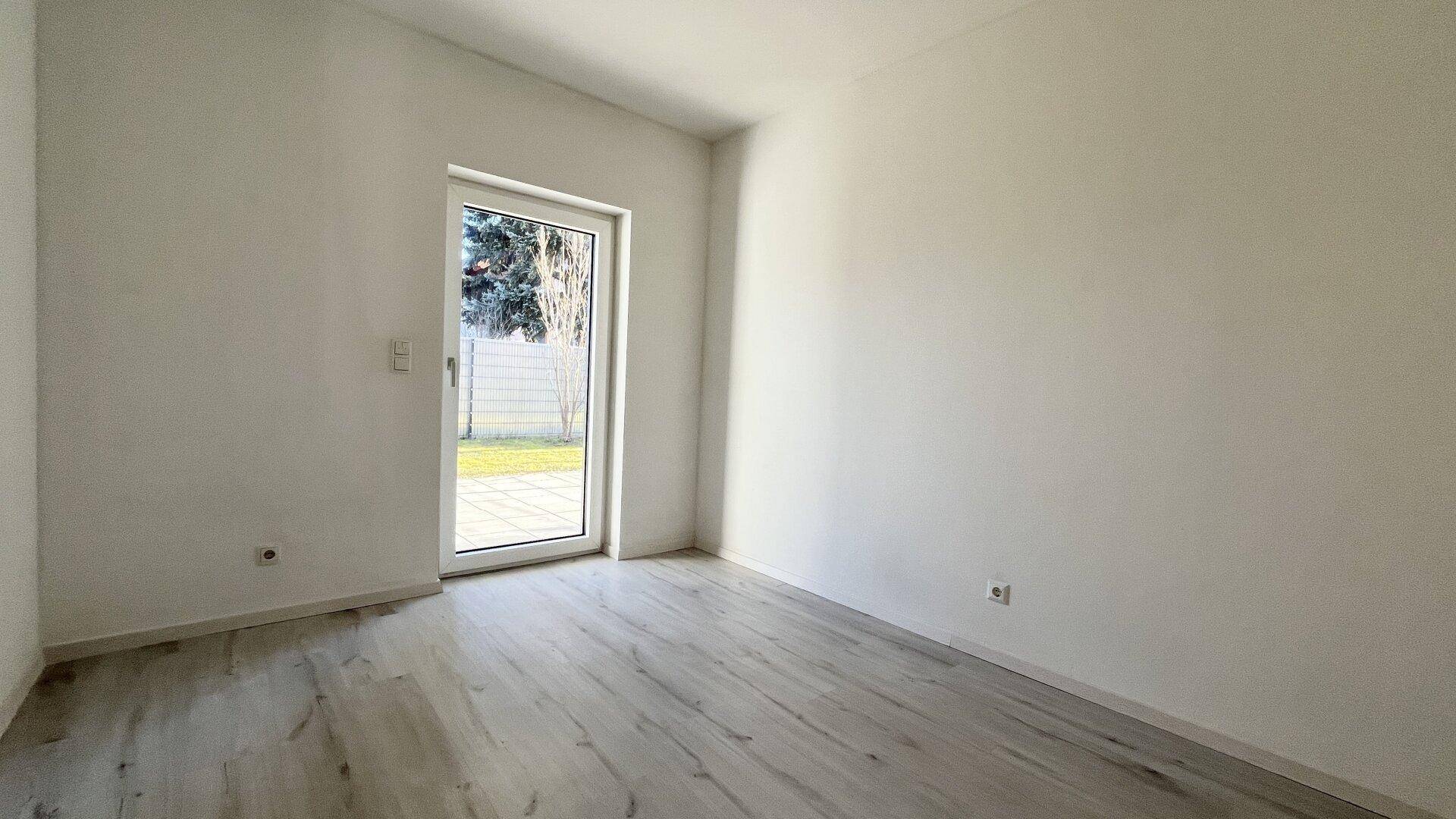 Leeres Zimmer mit Laminatboden und einer Tür, die ins Grüne führt.