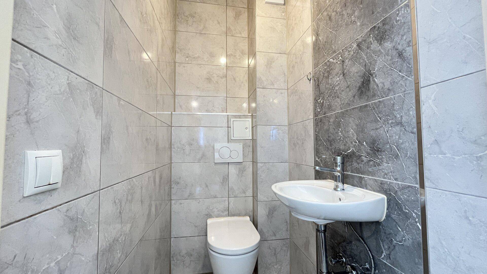 Modernes Gäste-WC mit Wand-WC, kleinem Waschbecken und grauen Fliesen.