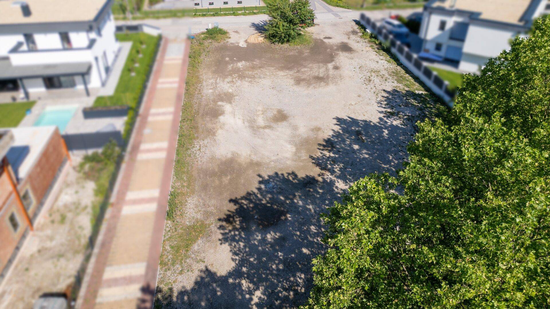 Luftaufnahme des unbebauten Grundstücks mit Schattenwurf von Bäumen und angrenzenden Wegen und Häusern.