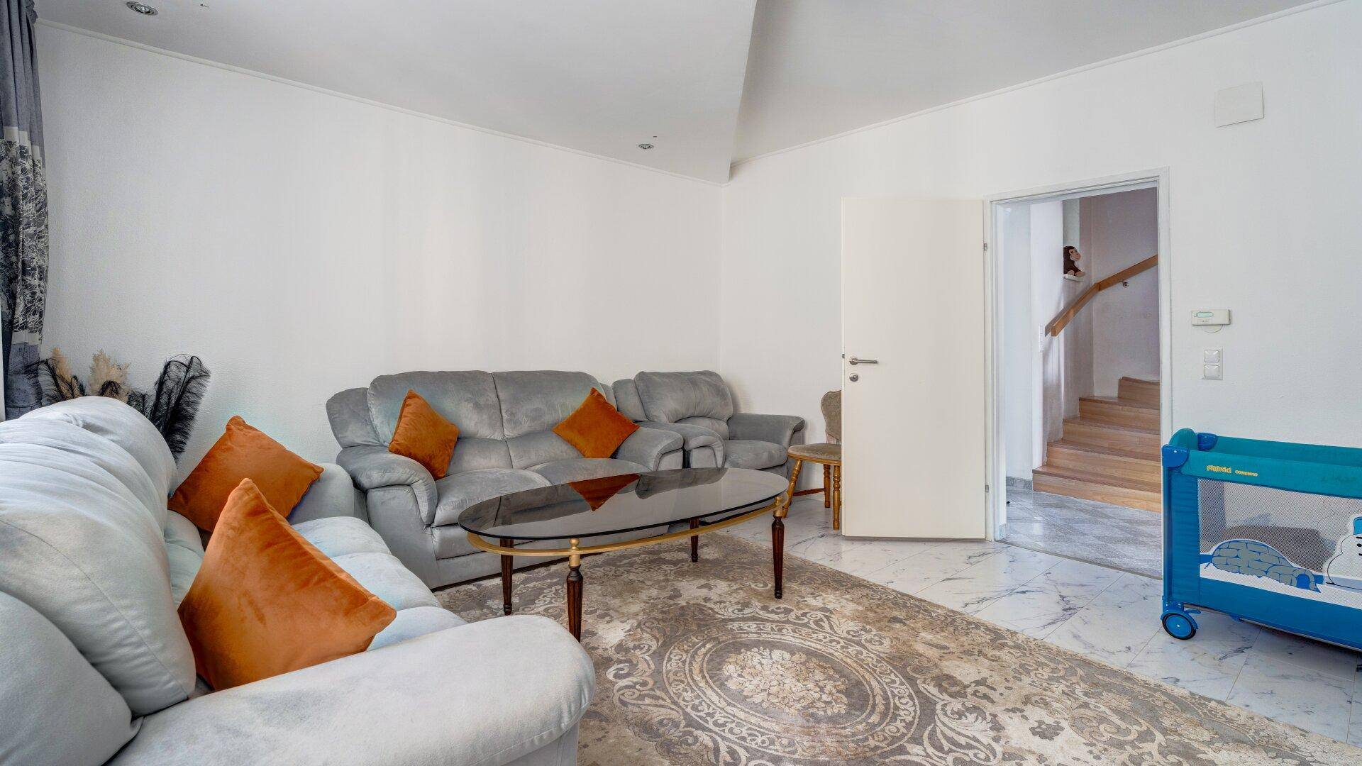 Wohnzimmer mit grauen Sofas, einem Babybett und Zugang zu einer Holztreppe.