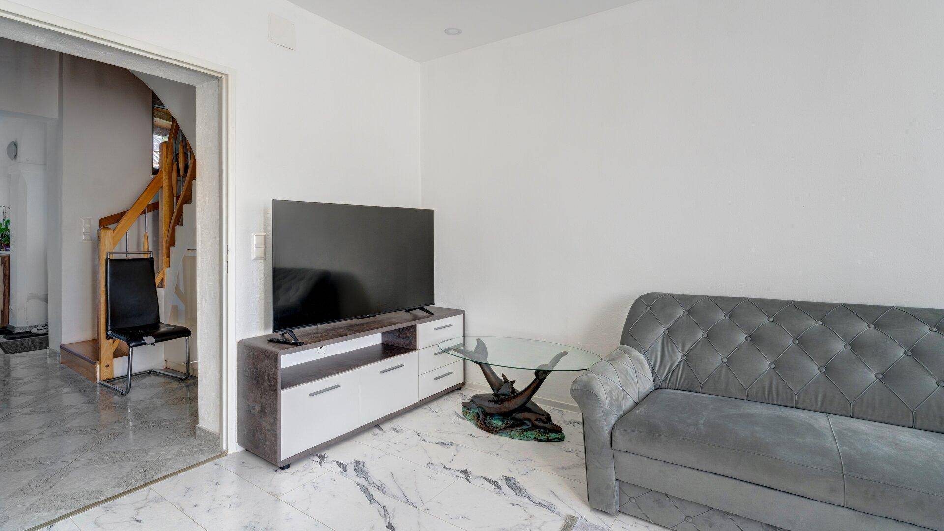 Wohnzimmerecke mit Fernseher, Couchtisch und Blick auf den Flur mit der Holztreppe.