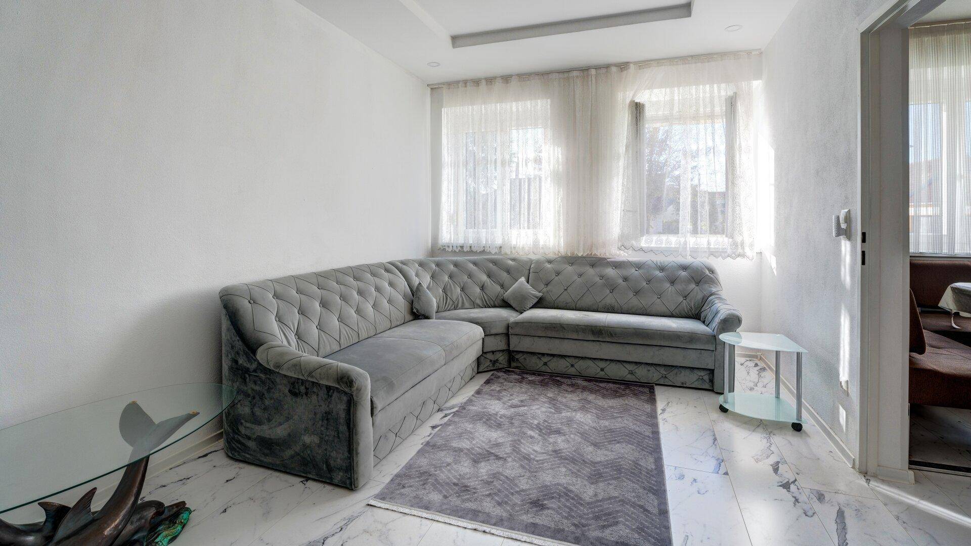 Helles Wohnzimmer mit großem Ecksofa, kleinen Beistelltischen und transparenten Vorhängen.