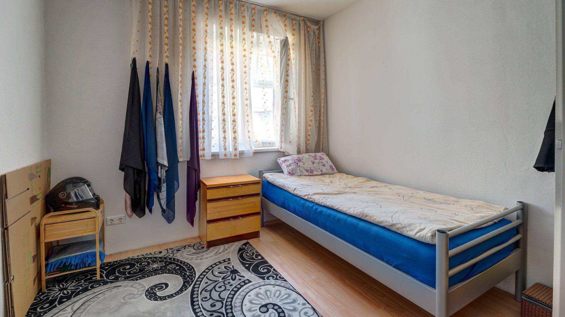 Kleines Schlafzimmer mit Einzelbett, Nachttisch und einem Fenster mit Vorhängen.