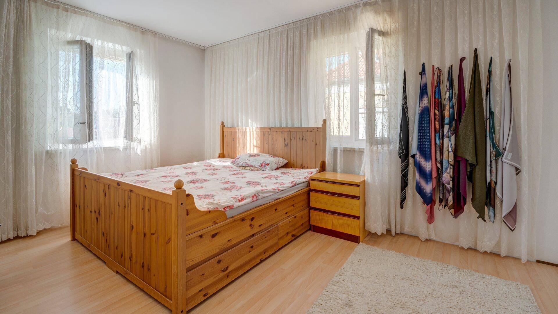 Helles Schlafzimmer mit Doppelbett aus Holz, zwei Fenstern und transparenten Vorhängen.