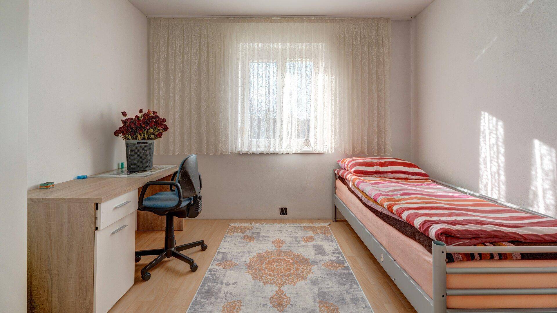 Helles Schlafzimmer mit Einzelbett, Schreibtisch und Fenster, ausgestattet mit einem Teppich.