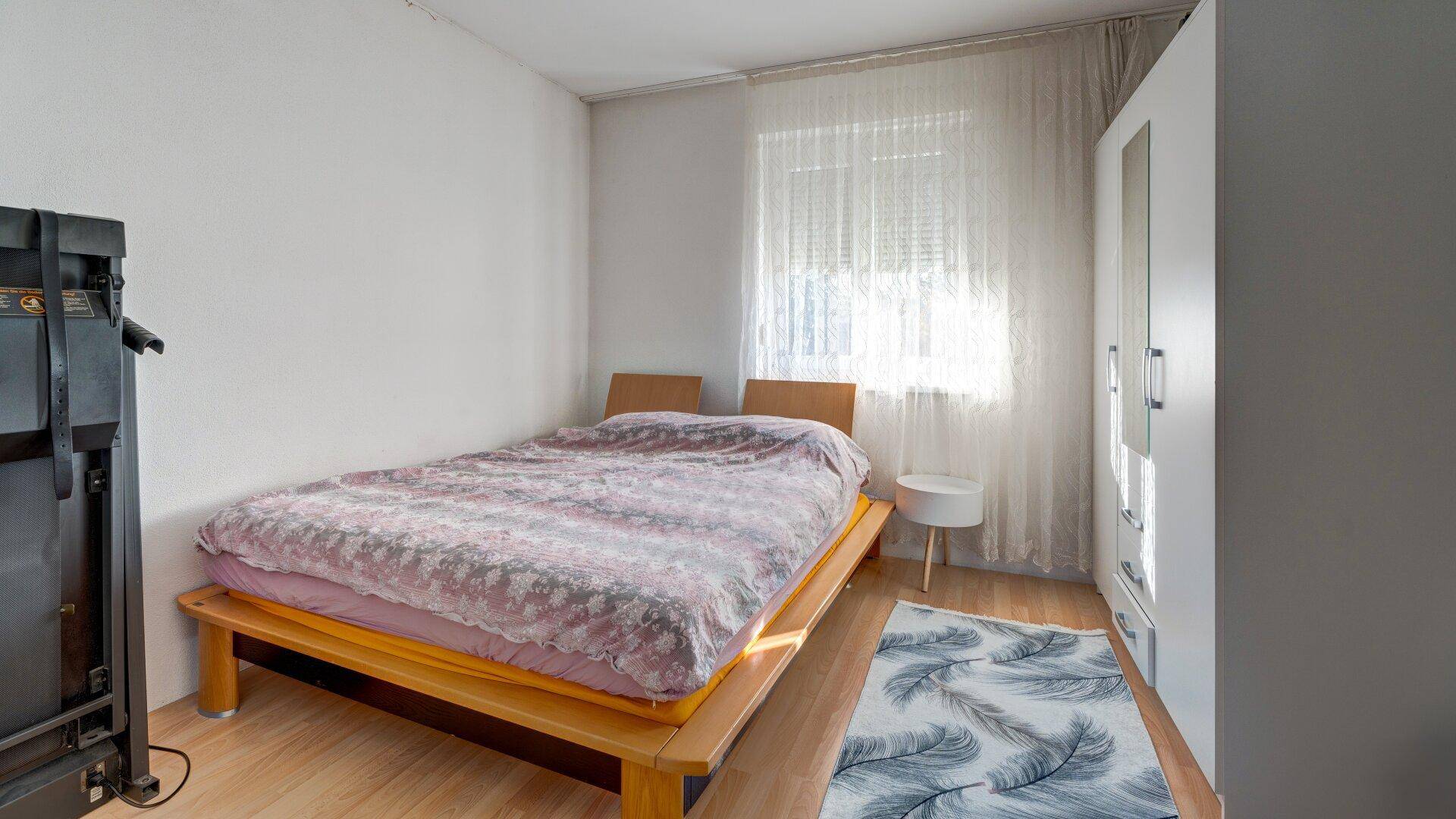 Schlafzimmer mit Doppelbett, kleinem Nachttisch und Kleiderschrank, hell durch das Fenster.