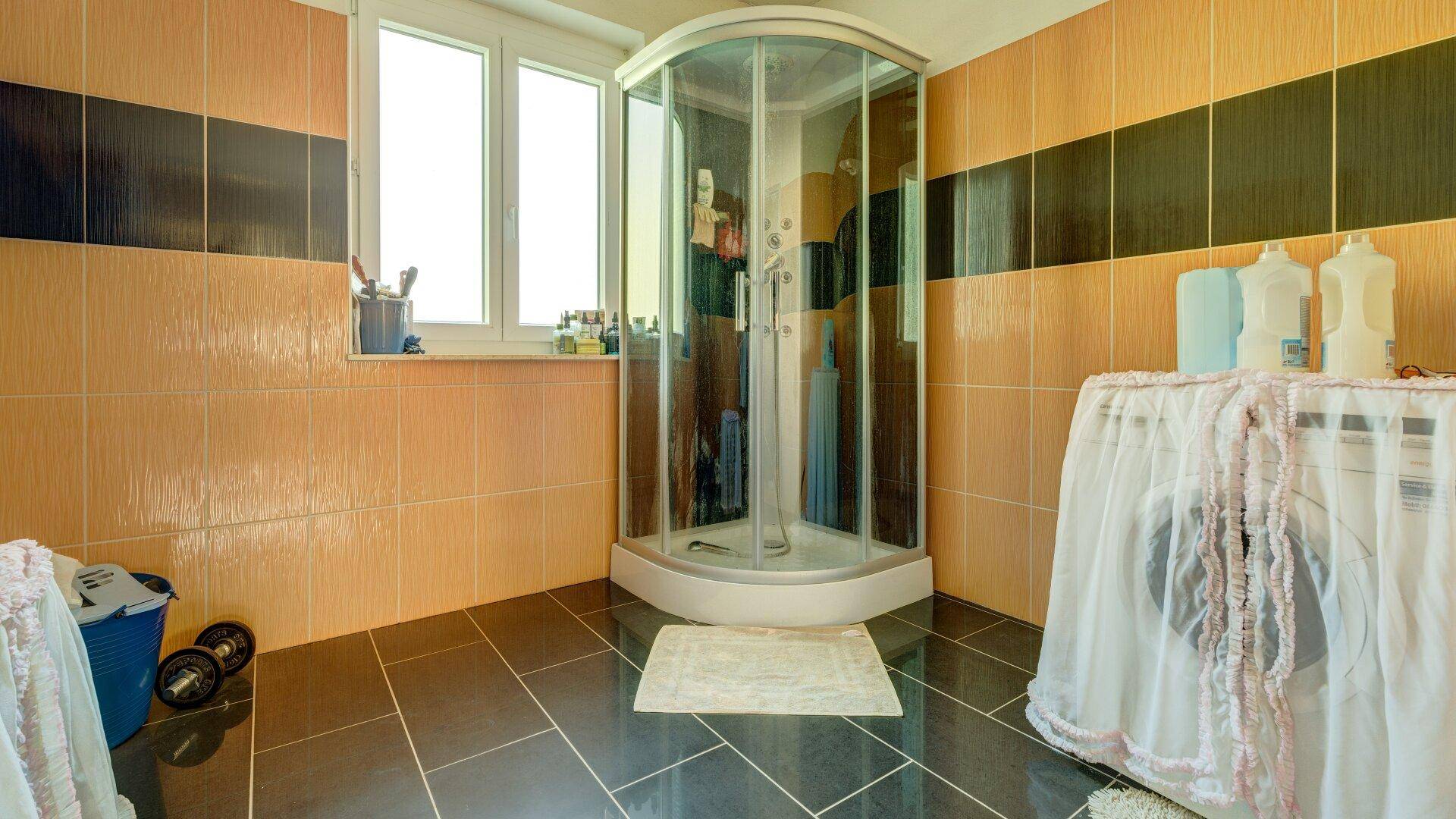 Badezimmer mit orange-schwarzen Fliesen, Duschkabine, Fenster und Platz für eine Waschmaschine.
