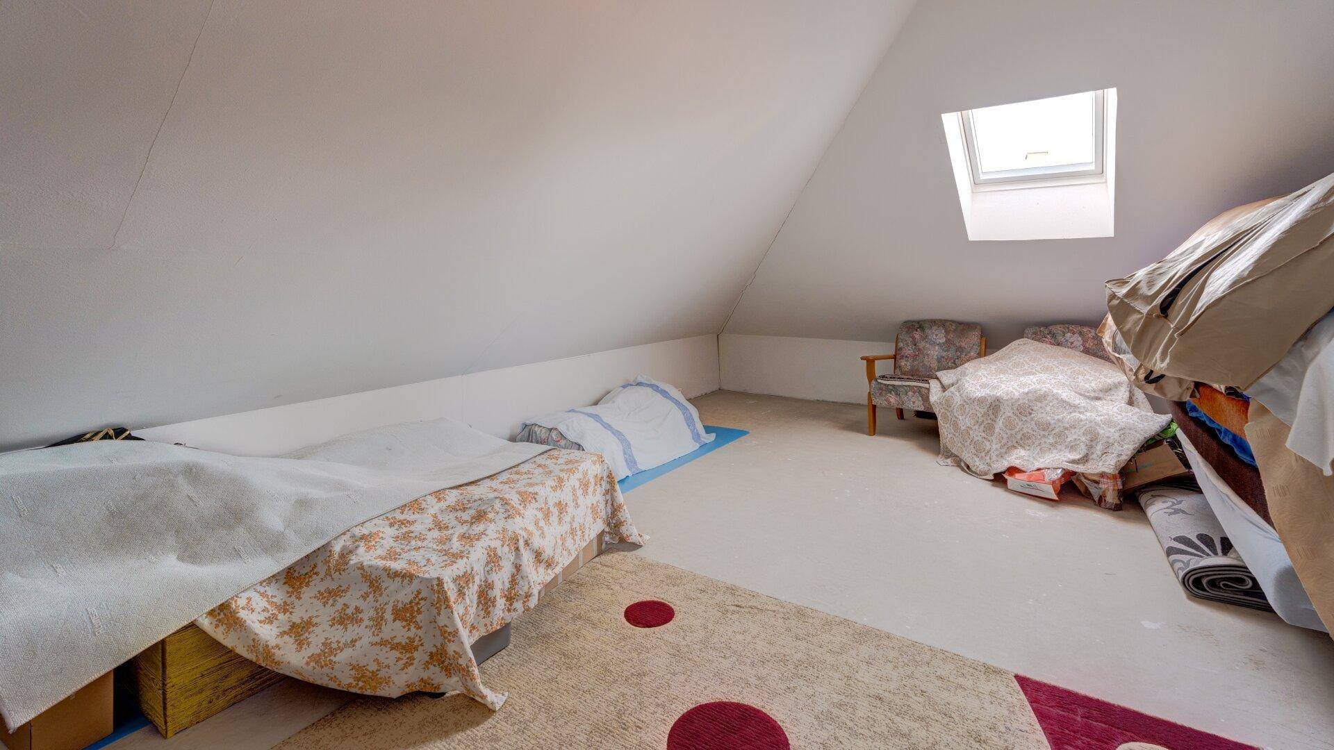 Dachzimmer mit schrägen Wänden, Dachfenster und provisorischen Schlafgelegenheiten oder Lagerflächen.