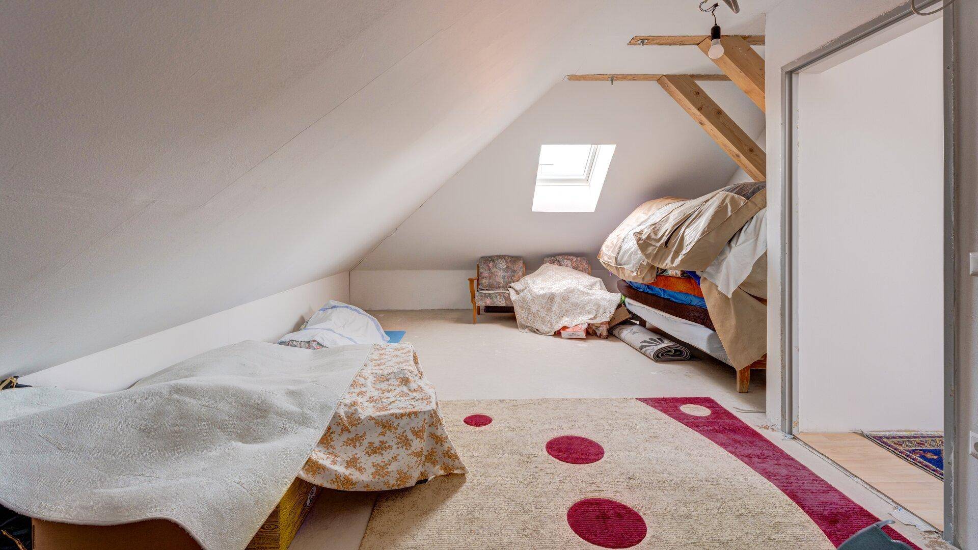 Helles Dachzimmer mit schrägen Wänden, Dachfenster und Zugang zu einem weiteren Bereich.