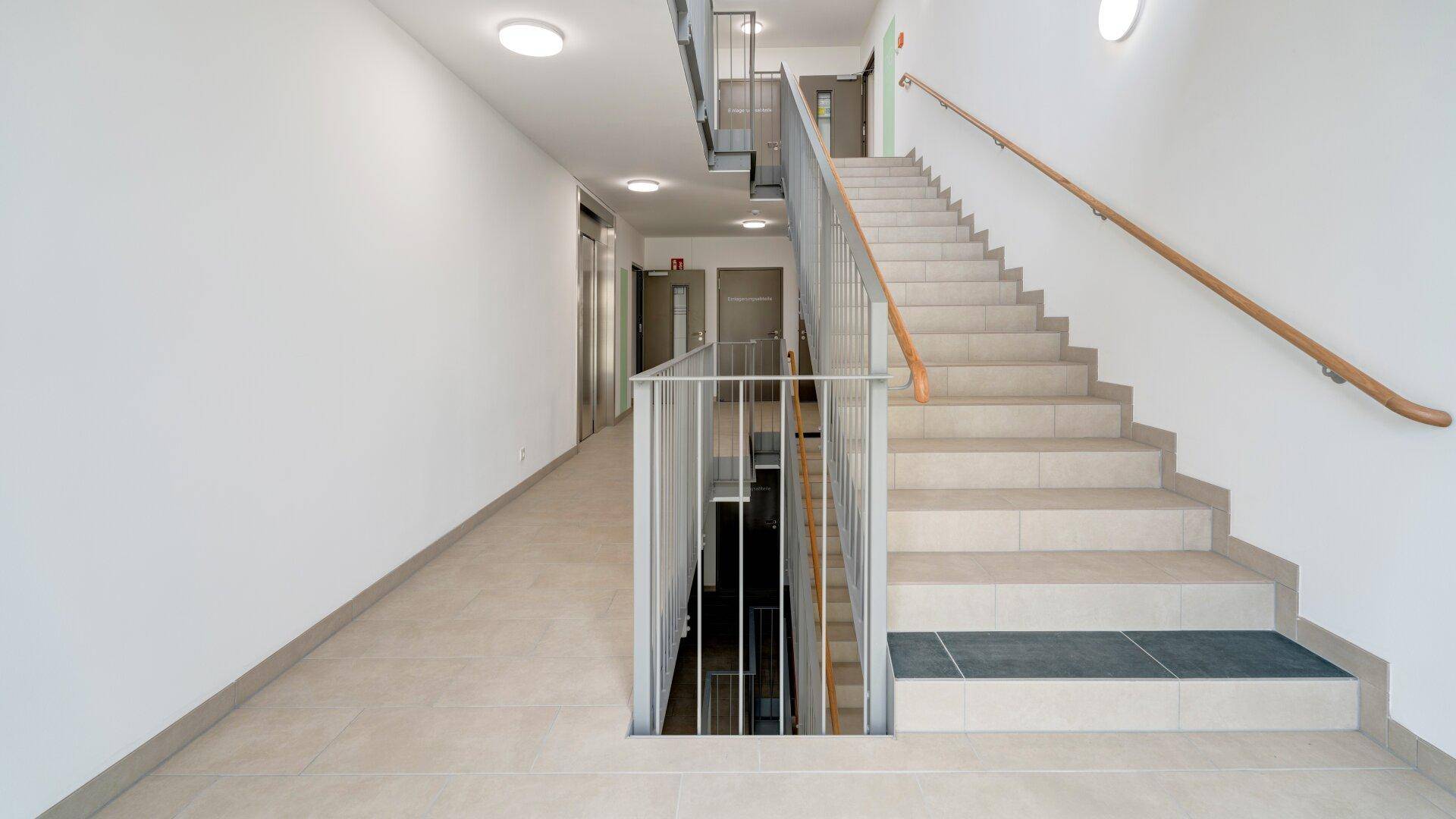 Geräumiges Treppenhaus mit Fliesenboden, Metallgeländer und Holzhandlauf.