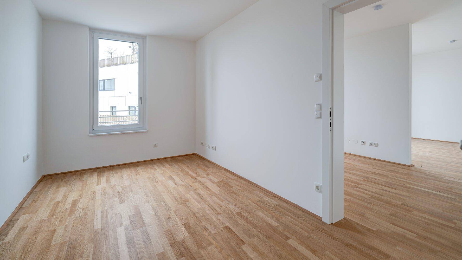 Leeres Zimmer mit Parkettboden und Fenster, das einen Blick nach draußen bietet.
