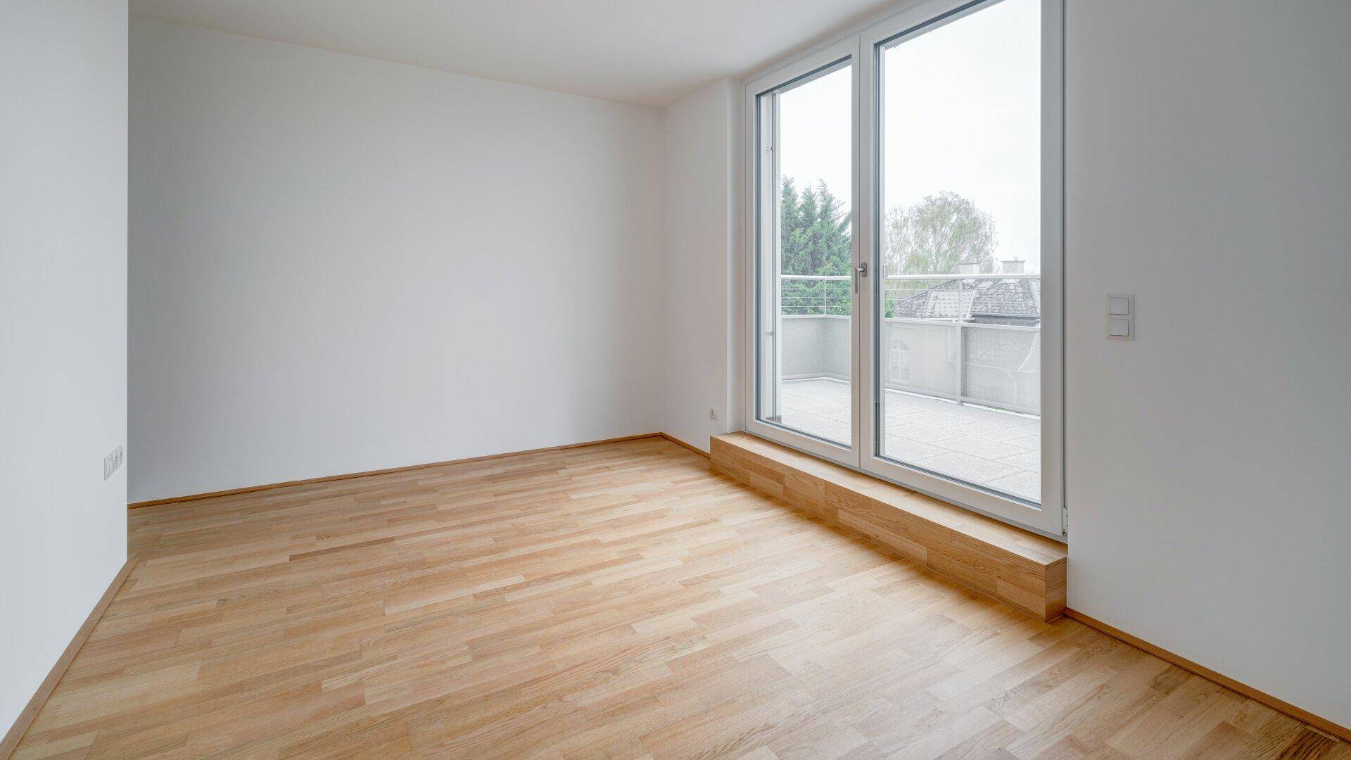 Heller, leerer Raum mit Parkettboden und großen Fenstern, die auf einen Balkon blicken.