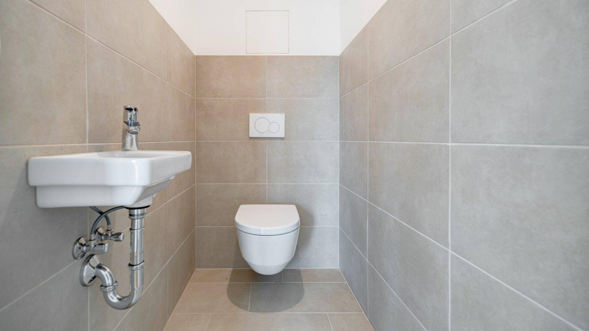 Modernes Gäste-WC mit wandhängender Toilette, kleinem Waschbecken und hellgrauen Fliesen.