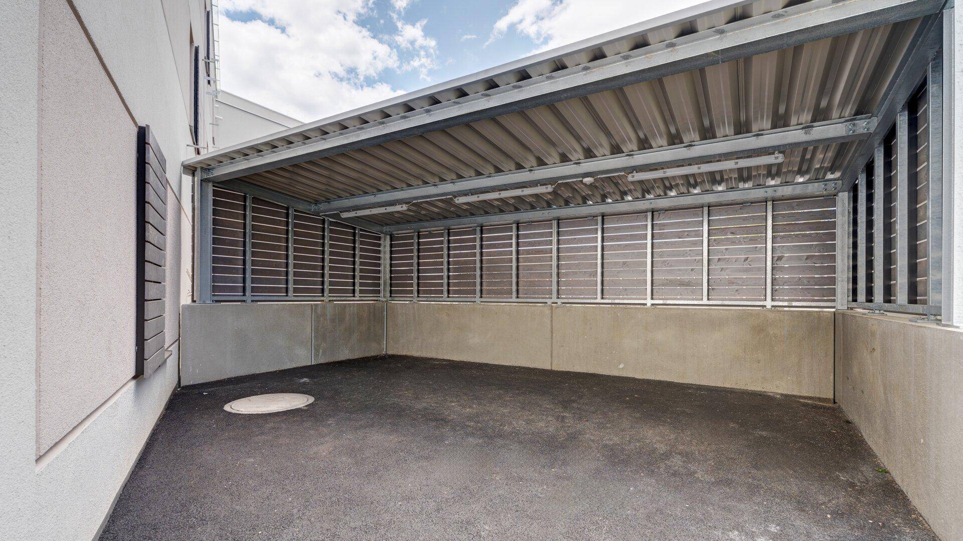 Überdachter Parkplatz mit Metallkonstruktion und Holzlamellenwänden für Fahrzeuge.