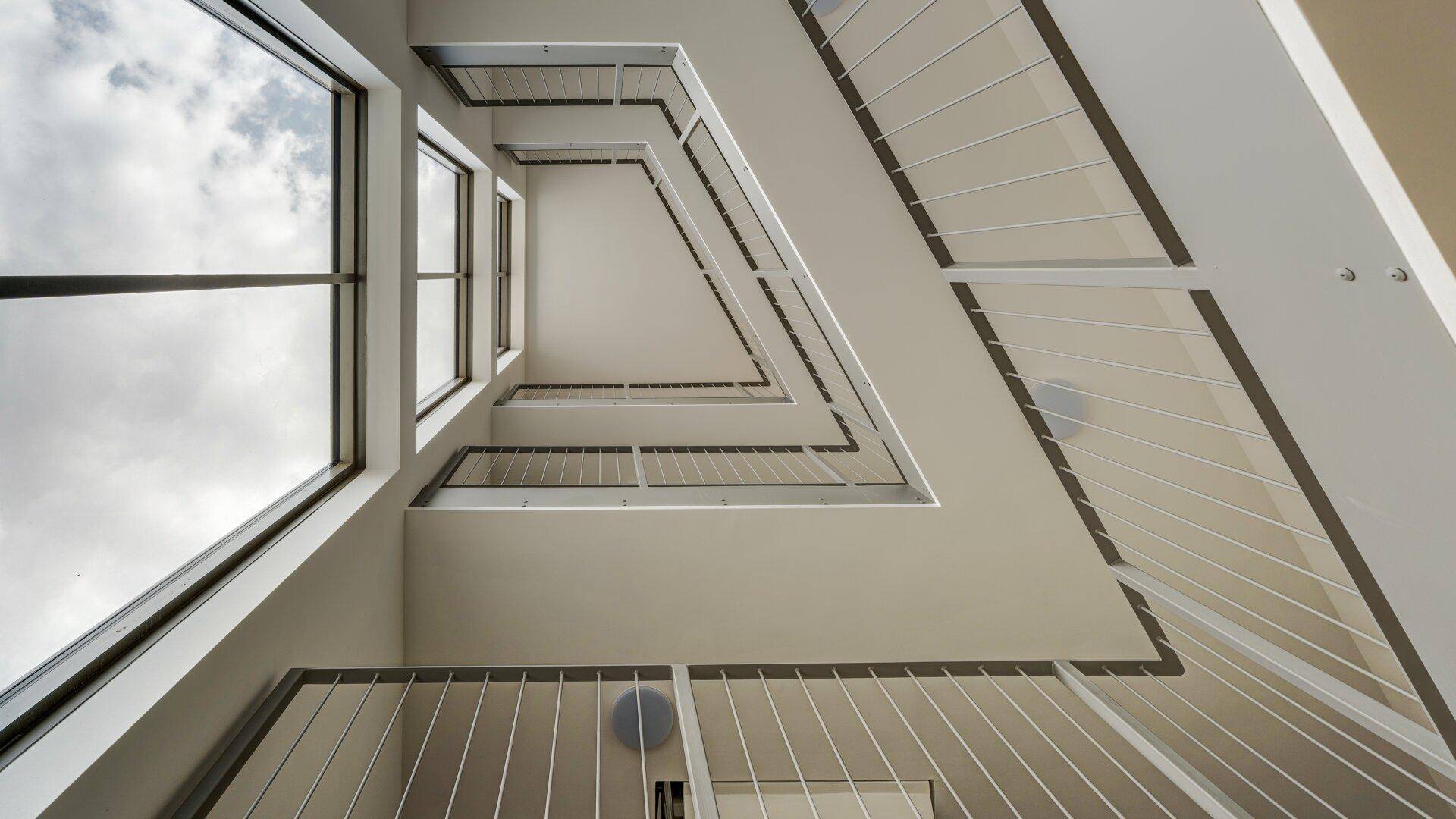 Blick nach oben in ein modernes Treppenhaus mit großen Fenstern und viel Tageslicht.