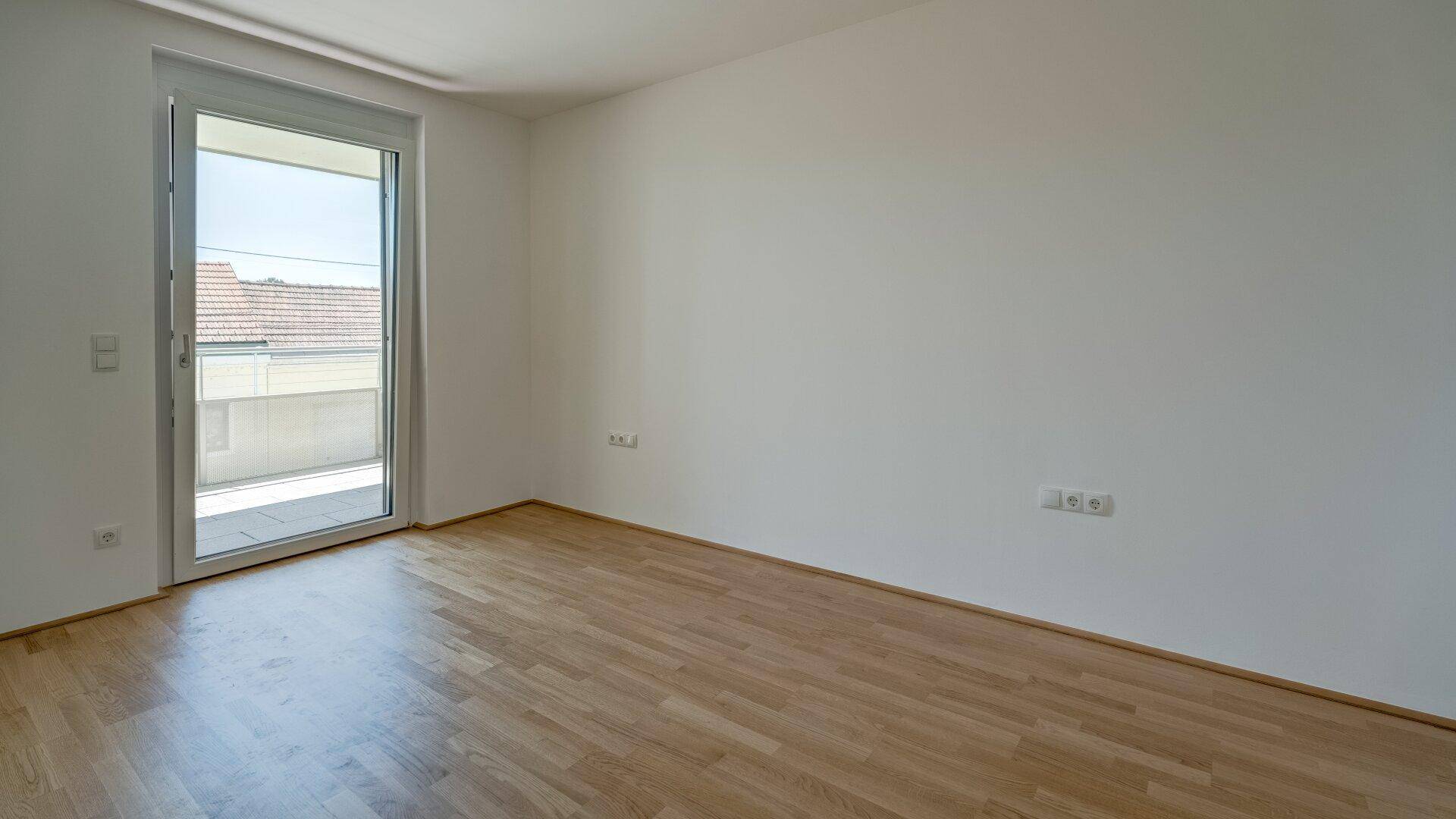 Schlafzimmer mit Parkettboden, weißen Wänden und direktem Zugang zum Balkon.