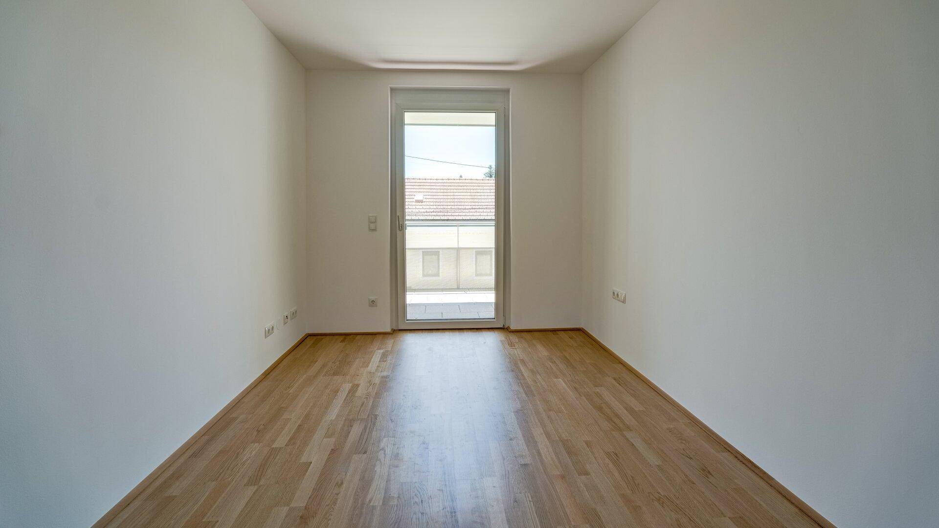 Helles Zimmer mit Parkettboden und großer Balkontür, die viel Licht hereinlässt.