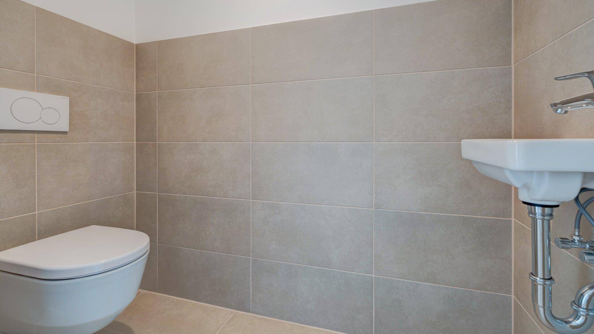Separates WC mit beigefarbenen Fliesen, wandhängender Toilette und kleinem Handwaschbecken.