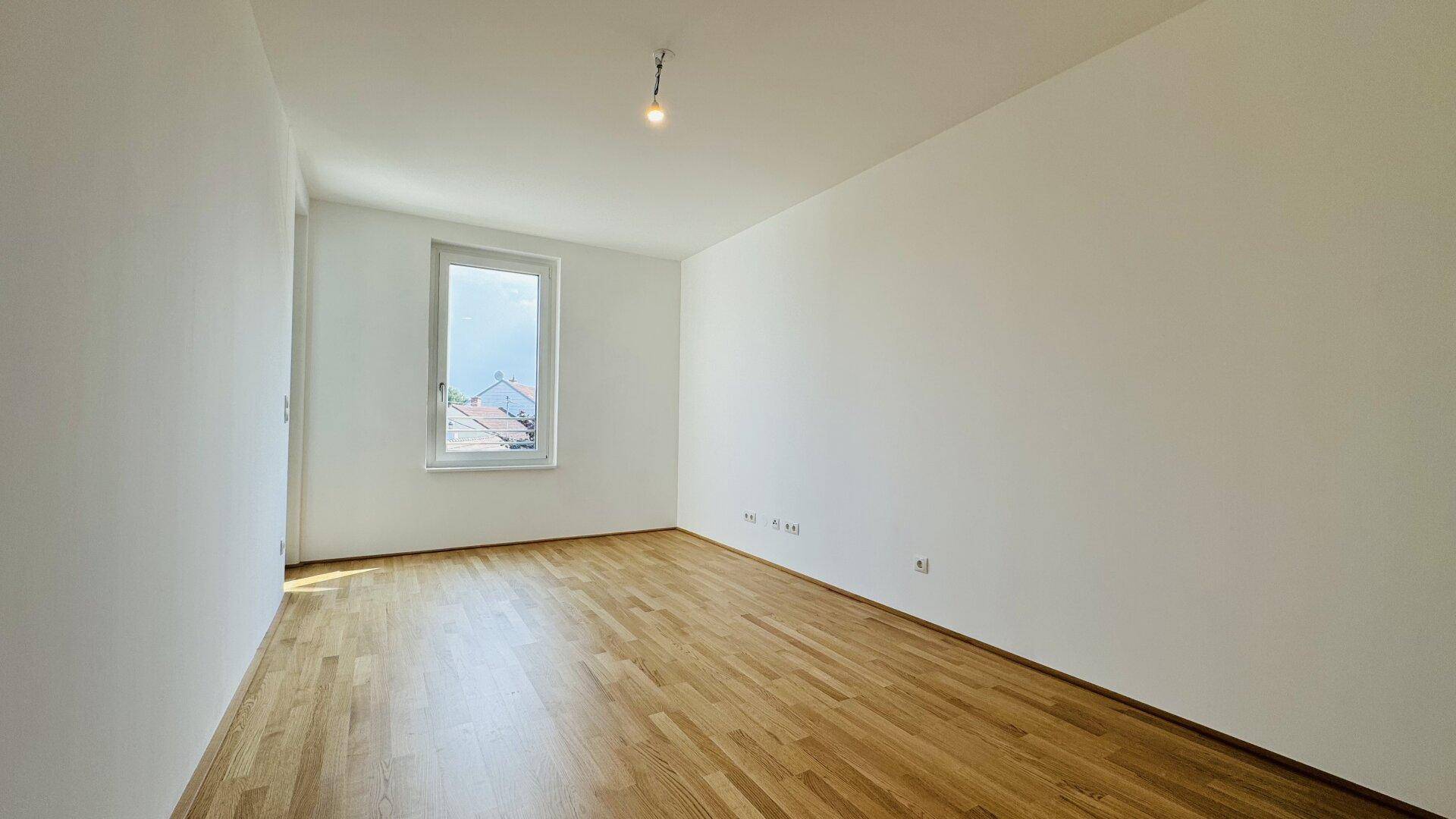 Leeres Zimmer mit Holzparkettboden und einem Fenster mit Blick ins Grüne.