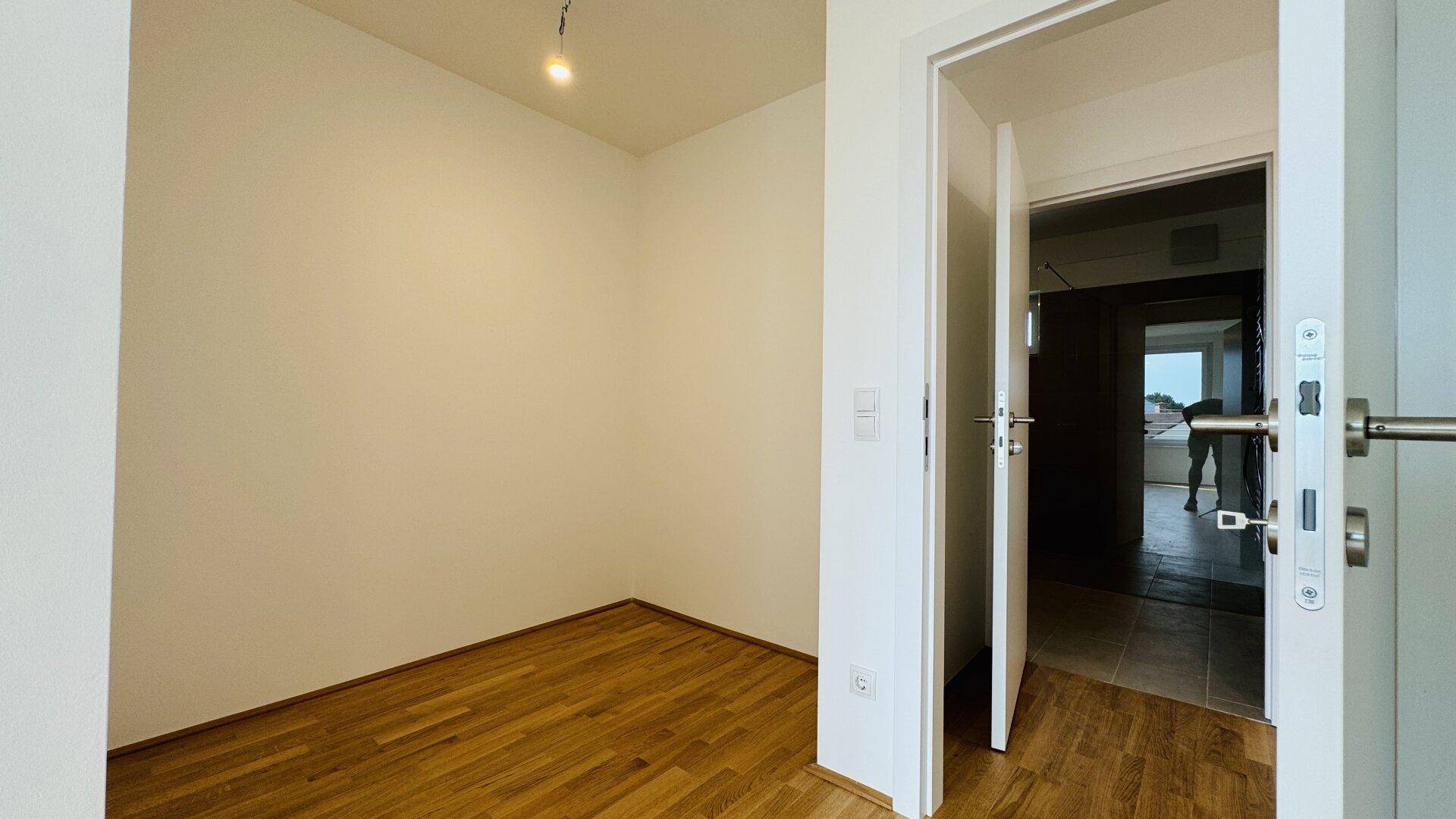 Kleiner, heller Raum mit Holzparkettboden und offener Tür zu einem weiteren Bereich.