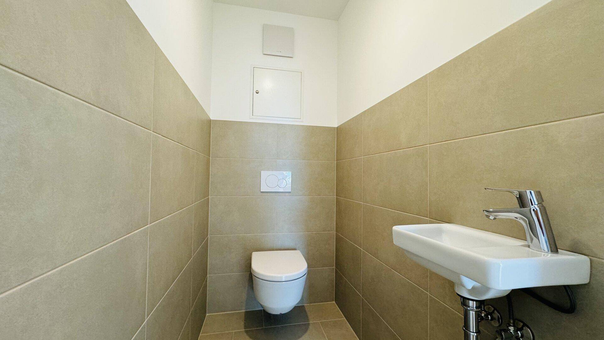 Gäste-WC mit wandhängender Toilette, kleinem Waschbecken und beigefarbenen Fliesen.