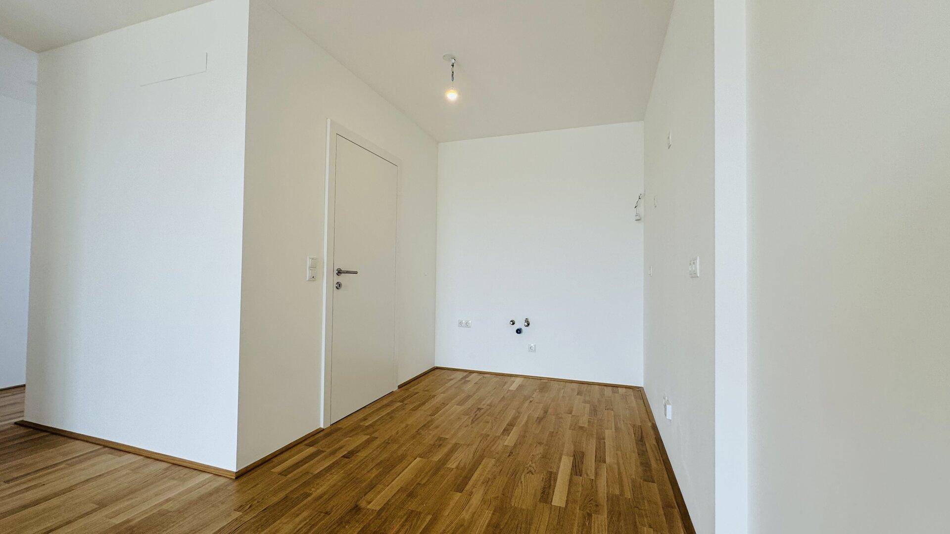 Leerer Raum mit Holzparkettboden und Anschlüssen an der Wand, möglicherweise für eine Küche.