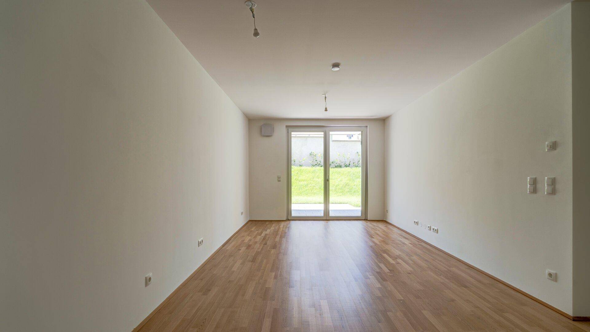 Helles, leeres Wohnzimmer mit Holzboden und Zugang zu einer Terrasse oder Garten.