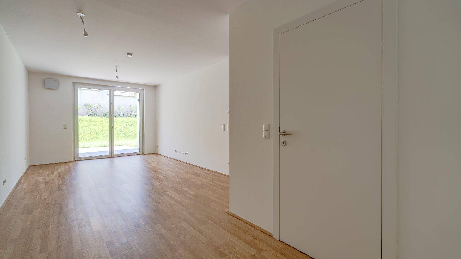 Geräumiges, unmöbliertes Wohnzimmer mit Parkettboden und weißer Tür in einem Neubau.