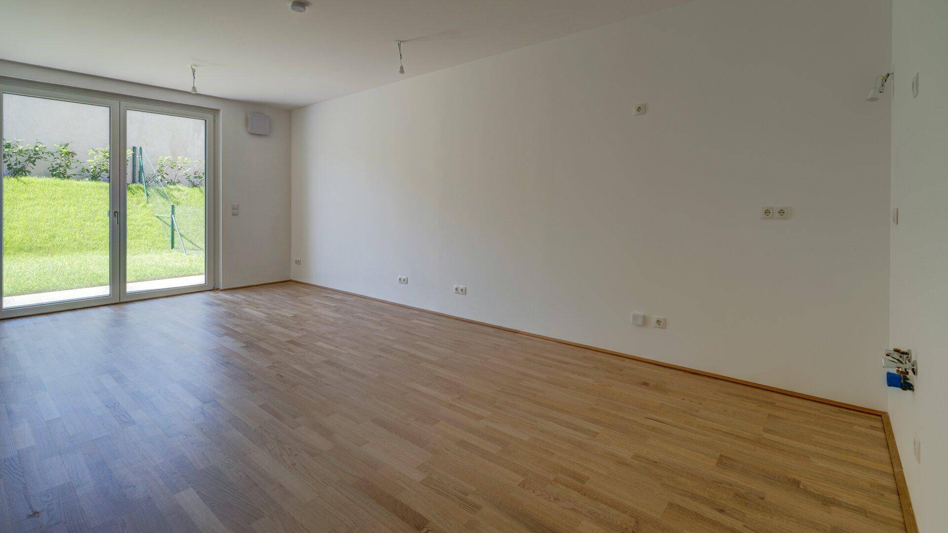 Heller, leerer Raum mit Holzboden und großen Fenstern, die den Blick in den Garten freigeben.