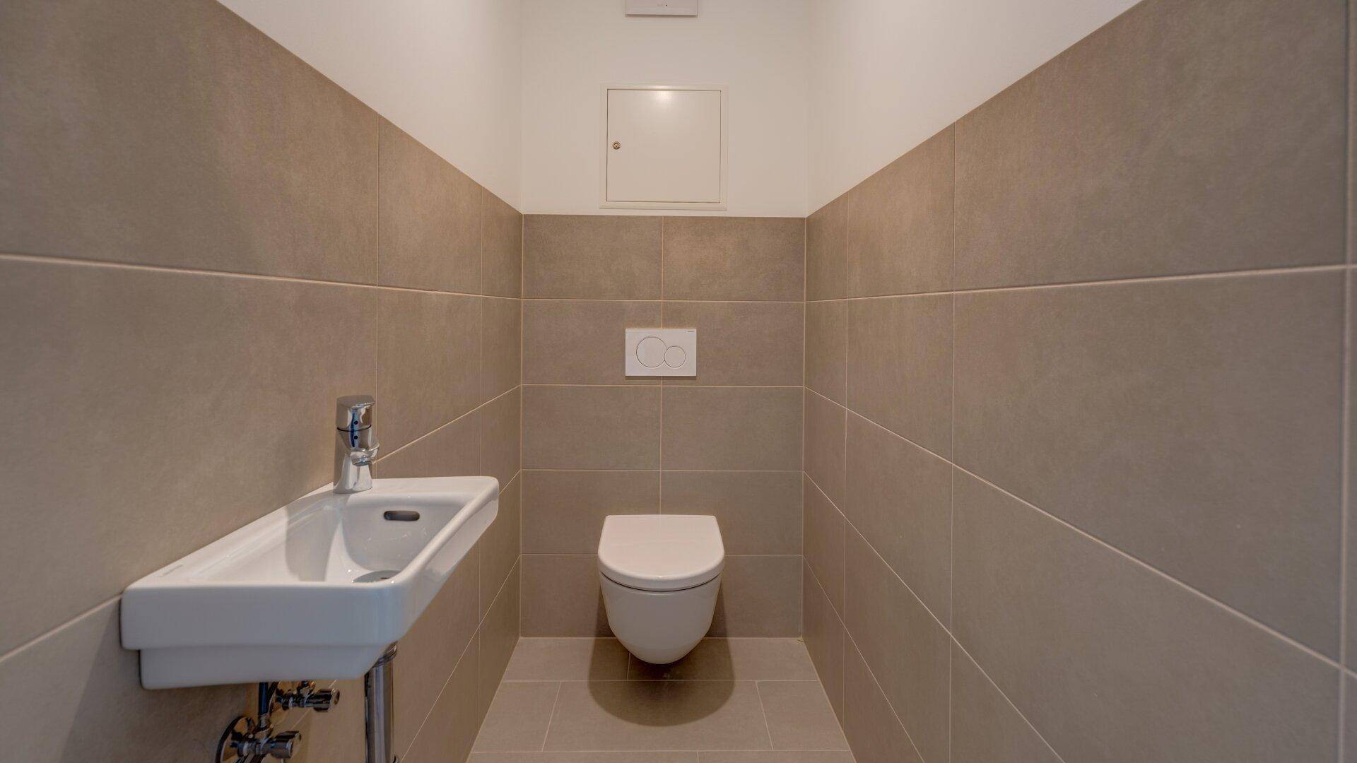 Modernes Gäste-WC mit beigefarbenen Fliesen, wandhängendem WC und kleinem Waschbecken.